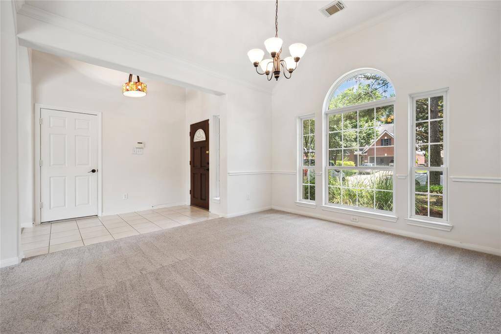 This room features a spacious, carpeted area with abundant natural light from large arched windows. It has a high ceiling with a stylish chandelier and a view of the well-maintained front yard. The entryway includes tiled flooring and a classic wooden front door.
