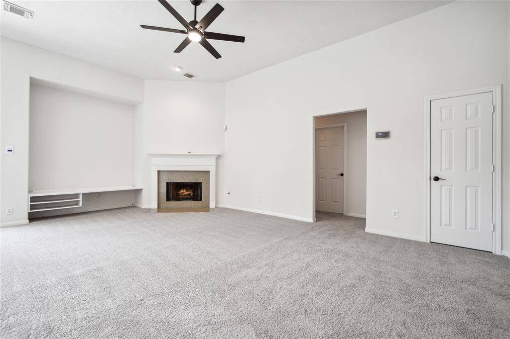 Spacious living room with neutral tones, featuring a cozy fireplace, built-in shelving, and a ceiling fan. The room has clean carpeting and ample natural light, creating a warm, inviting atmosphere.
