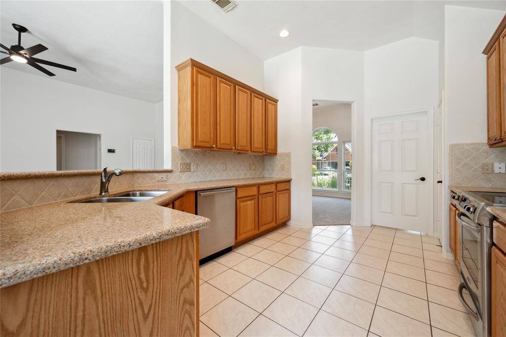 This kitchen features granite countertops, stainless steel appliances, and wooden cabinetry. The open layout leads to a bright living area with large windows, and tile flooring is present throughout. A ceiling fan and neutral tones add a modern touch.