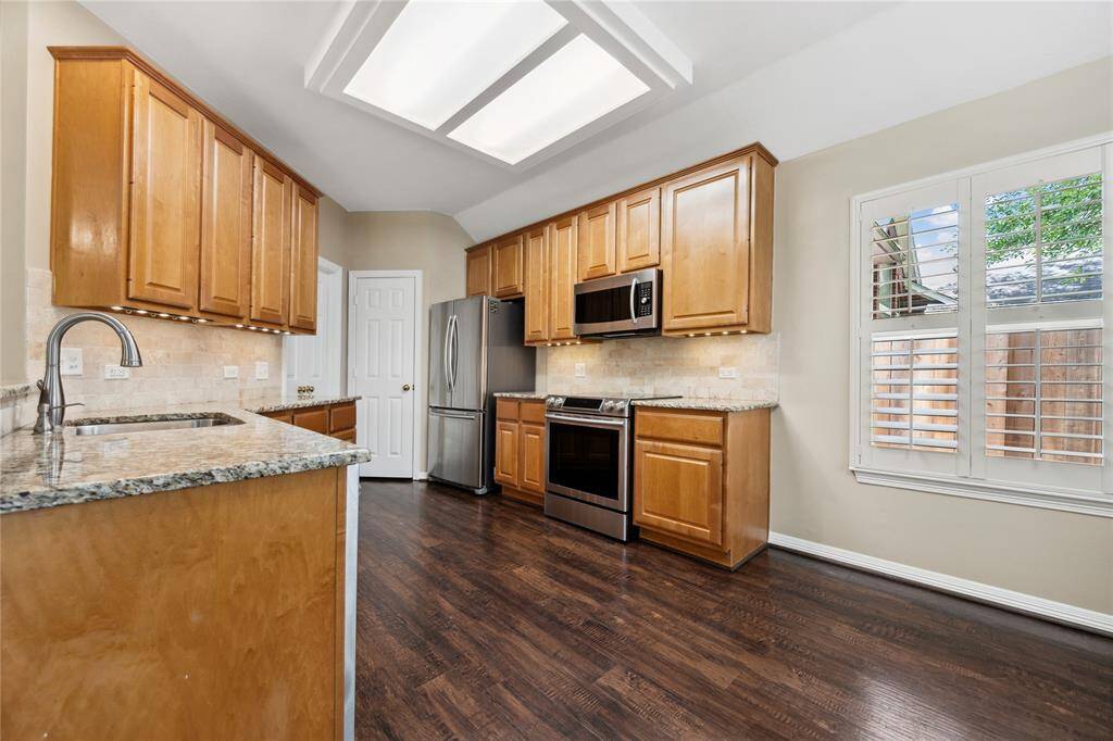 This kitchen features light wood cabinetry, granite countertops, and stainless steel appliances. It has dark hardwood floors and a bright window with shutters, creating a warm and inviting space.