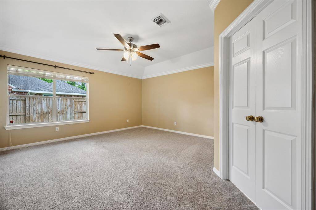 This spacious room features soft carpet flooring, neutral beige walls, and a ceiling fan with lighting. A large window allows natural light, and double doors add a touch of elegance, making it ideal for a bedroom or office space.