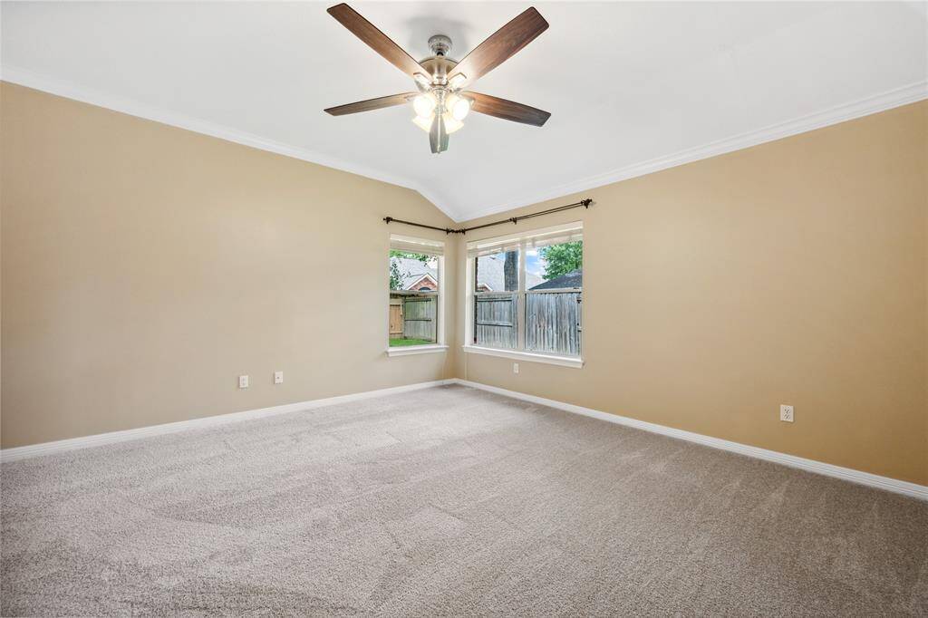 This spacious primary bedroom features neutral walls, plush carpeting, a ceiling fan, and large windows that provide natural light and a view of the fenced backyard.