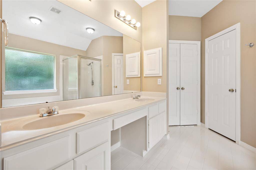 This bathroom features a spacious double vanity with ample storage, a large mirror, and a glass-enclosed shower. The room is well-lit with neutral walls and a frosted window for privacy.