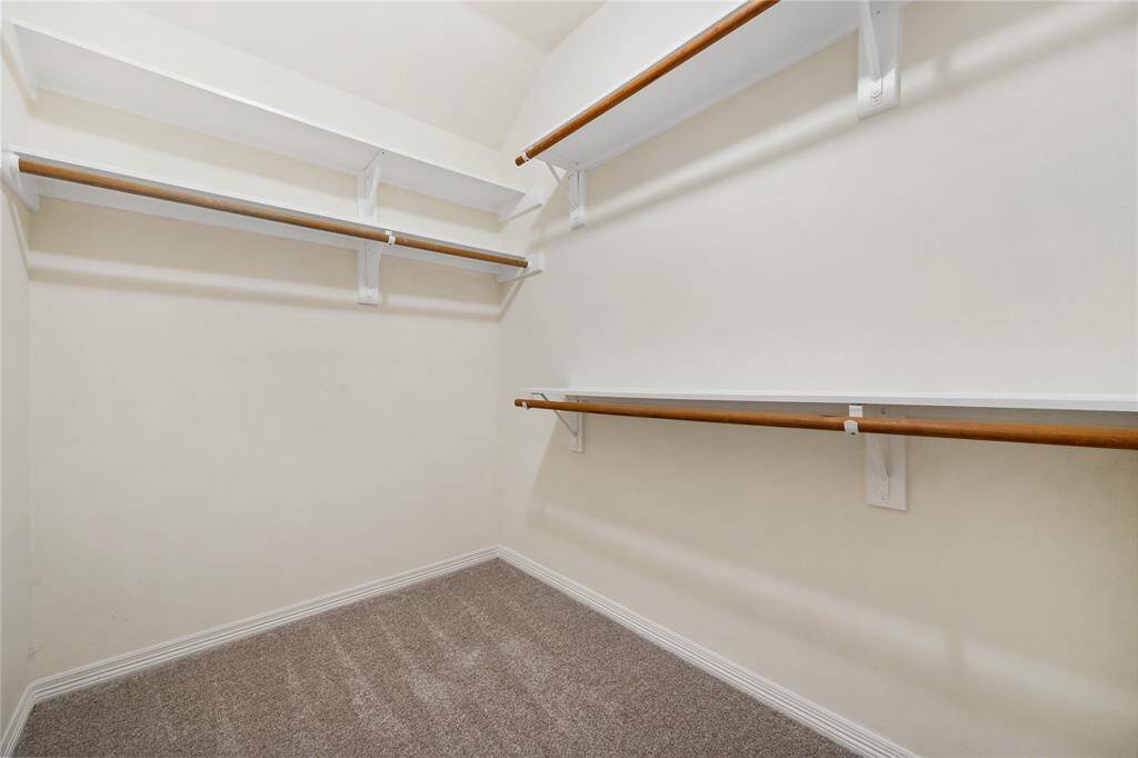 This photo shows a spacious walk-in closet with multiple wooden hanging rods and upper shelves, ideal for ample clothing storage. It features a neutral color palette and carpeted flooring.