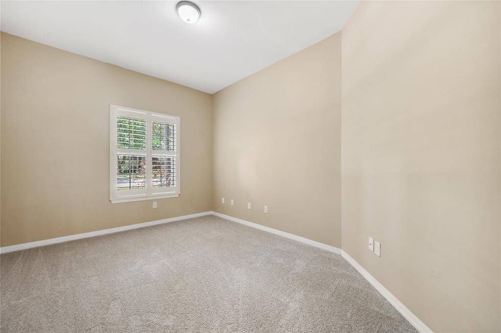 This photo shows a bright, empty room with beige walls and soft carpeting. There's a window with white shutters allowing natural light in, and a ceiling light fixture provides additional illumination. The space is clean and ready for customization.