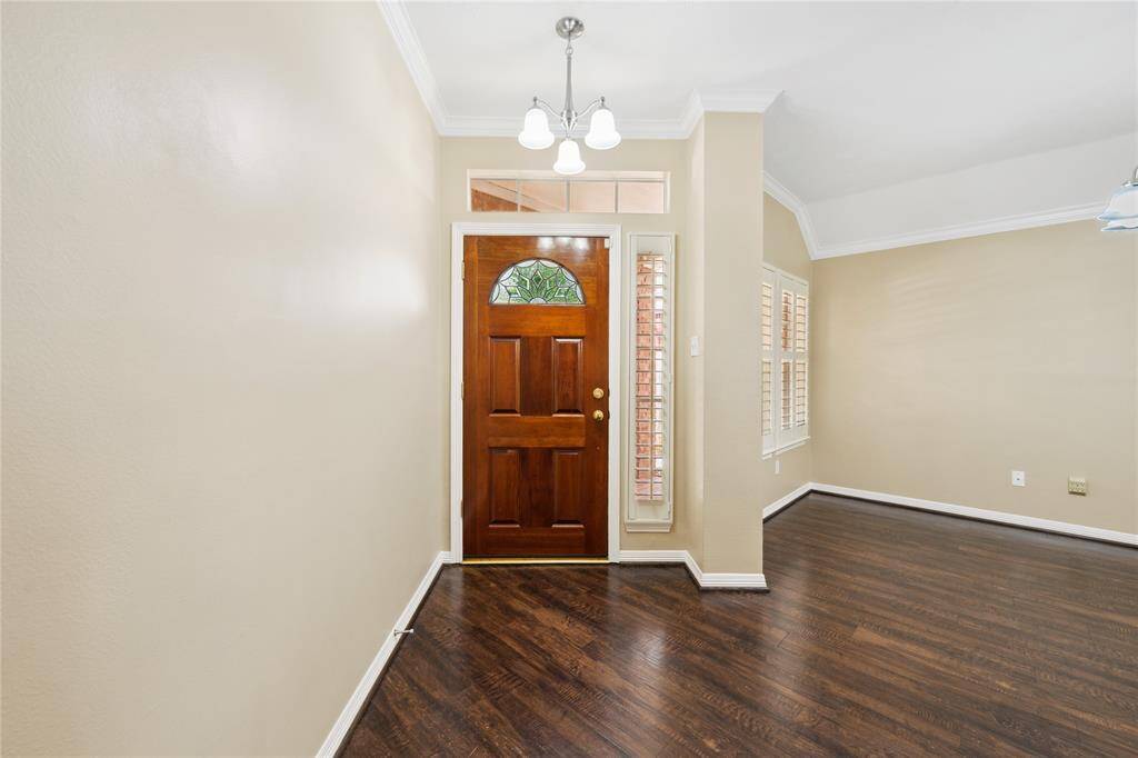 This entryway features a warm wooden door with decorative glass, complemented by light walls and dark hardwood flooring. Adjacent is a bright, airy room with large windows and crown molding, creating an inviting and elegant atmosphere.