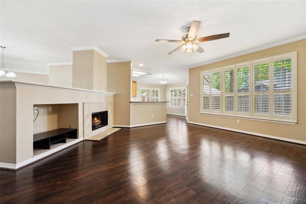 Bright and spacious living area with dark wood  floors, a cozy fireplace, and large windows with plantation shutters. The room features an open layout with a ceiling fan and access to the kitchen, creating an inviting and functional space.