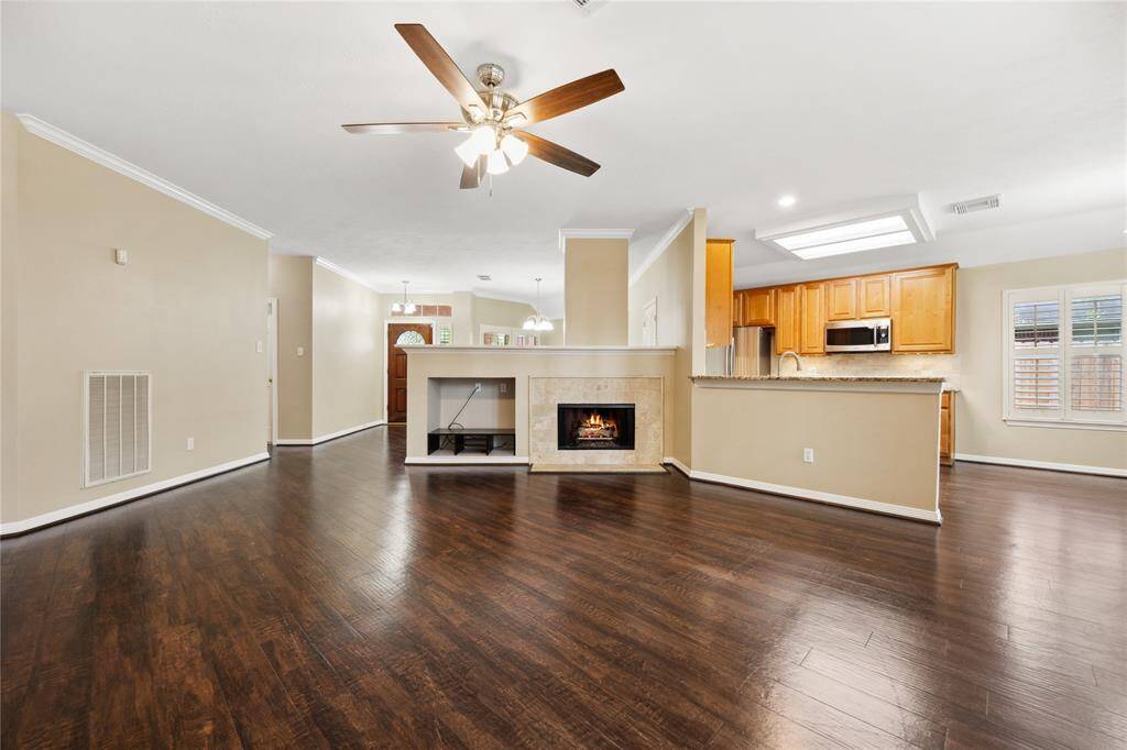 This spacious open-concept living area features rich wood floors, a cozy fireplace, and a modern kitchen with wooden cabinetry and stainless steel appliances. The room is bright and airy, enhanced by a ceiling fan and recessed lighting.