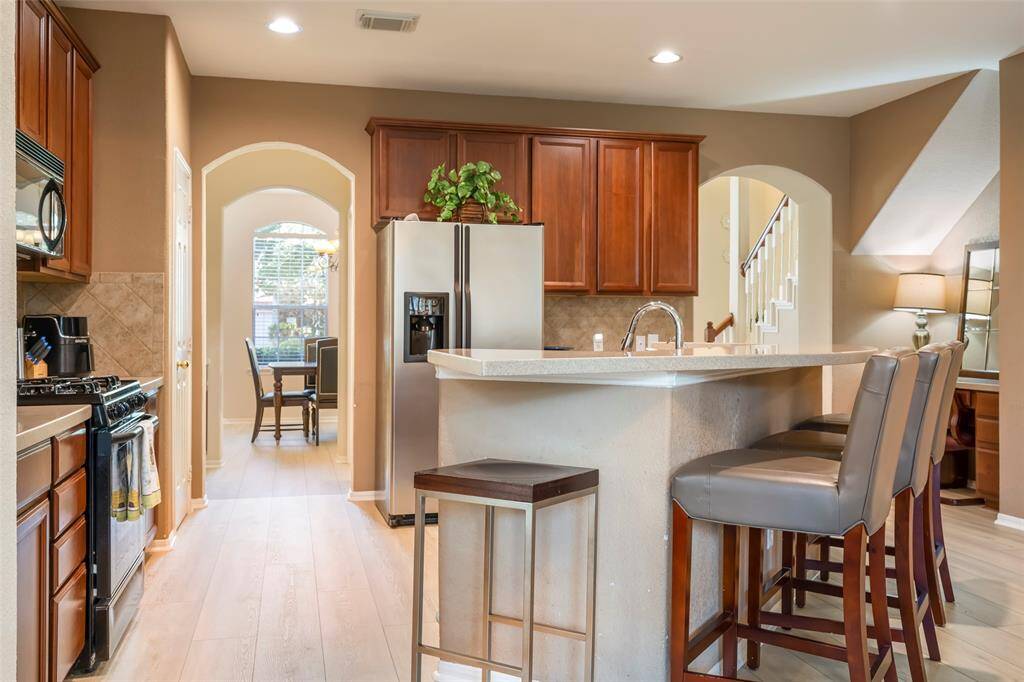 This inviting kitchen features warm wood cabinetry, a modern stainless steel refrigerator, and a cozy breakfast bar with seating. An arched doorway leads to a bright dining area, while soft lighting enhances the welcoming atmosphere.