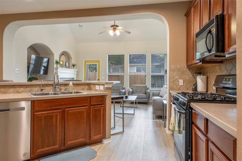 This photo showcases a cozy, open-concept kitchen and living area. The kitchen features warm wooden cabinetry, stainless steel appliances, and a gas stove. An arched opening connects to a bright living room with large windows, modern furniture, and a ceiling fan, creating a welcoming, airy space.