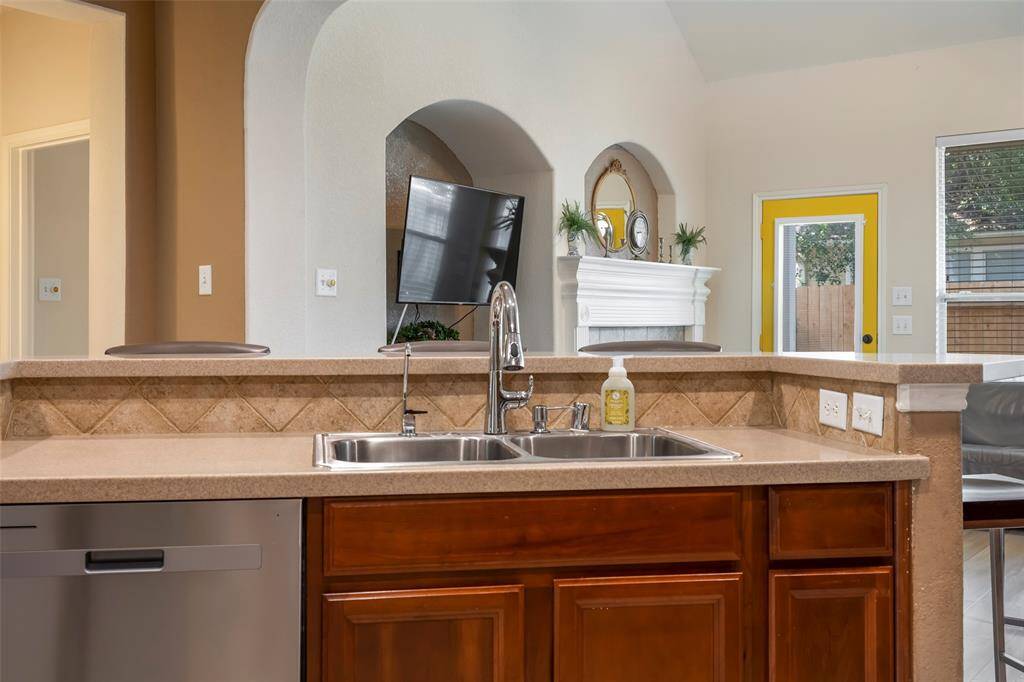 This photo showcases a kitchen with a dual sink, tiled backsplash, and a view into a cozy living area. There's a fireplace with a mantle and a large window letting in natural light. The space has a modern touch with a mounted TV and a vibrant yellow door.