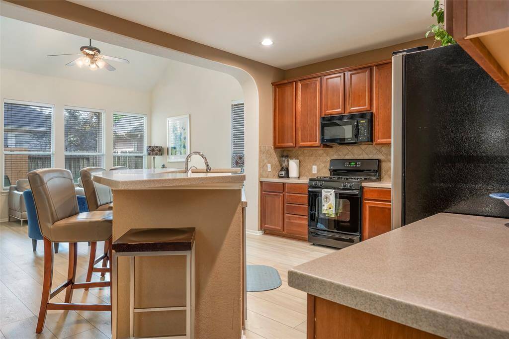 This inviting kitchen features warm wood cabinetry, modern black appliances, and a cozy breakfast bar with seating. It opens to a bright dining area with large windows, offering plenty of natural light and a view of the backyard.
