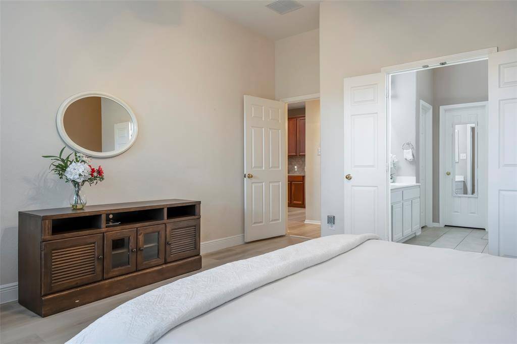 This bedroom features neutral tones, a mirror above a dark wood cabinet, and an en-suite bathroom with double doors, adding a touch of elegance and convenience.