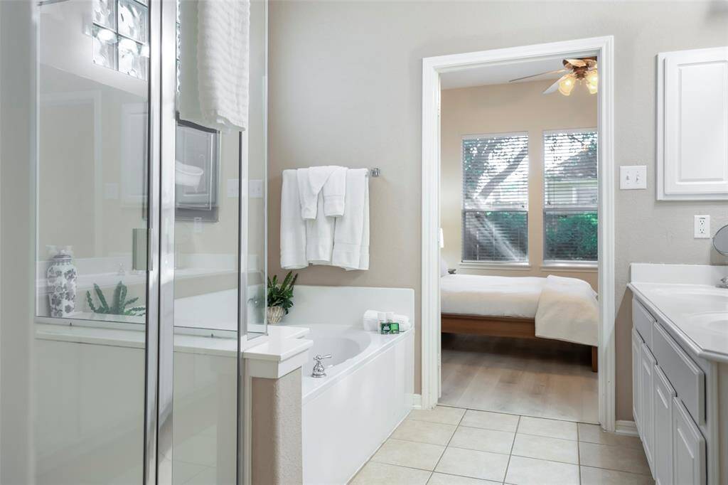 This photo showcases a bright bathroom with a glass-enclosed shower and a bathtub. It opens into a bedroom with large windows, allowing natural light to fill both spaces. The neutral tones create a calm and inviting atmosphere.