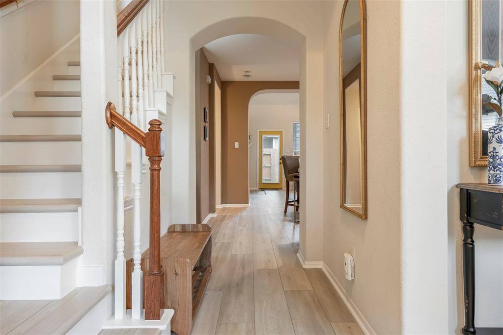 This inviting hallway features light wood flooring, a stylish staircase with white spindles and wood handrails, and a large mirror accentuating the space. The open design leads to a bright room in the background, creating a welcoming and airy atmosphere.