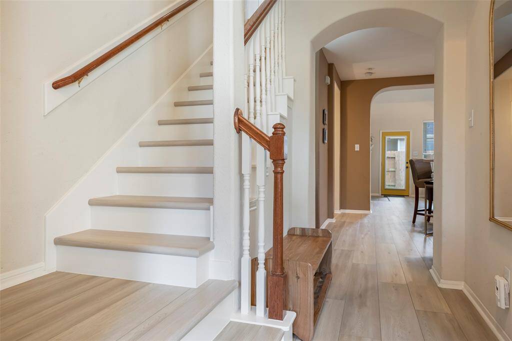 This photo showcases a welcoming entryway with light wood flooring and a staircase featuring wooden railings. The space is bright, with a hallway leading to a dining area and a door with a yellow frame. The neutral color palette enhances the airy feel.