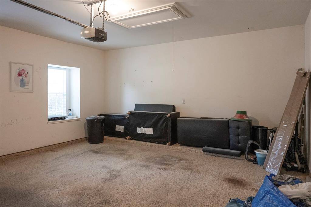 This photo shows a spacious garage with a window for natural light. It has a ceiling-mounted garage door opener and some stored items, offering potential for organization or additional storage.