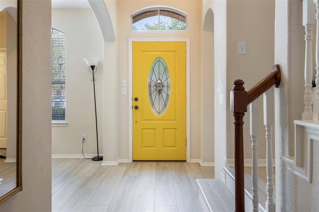 Bright entryway with a striking yellow door featuring an oval glass design. The area has light wood flooring, neutral walls, and a window for natural light. A staircase with wooden handrails adds a touch of elegance.