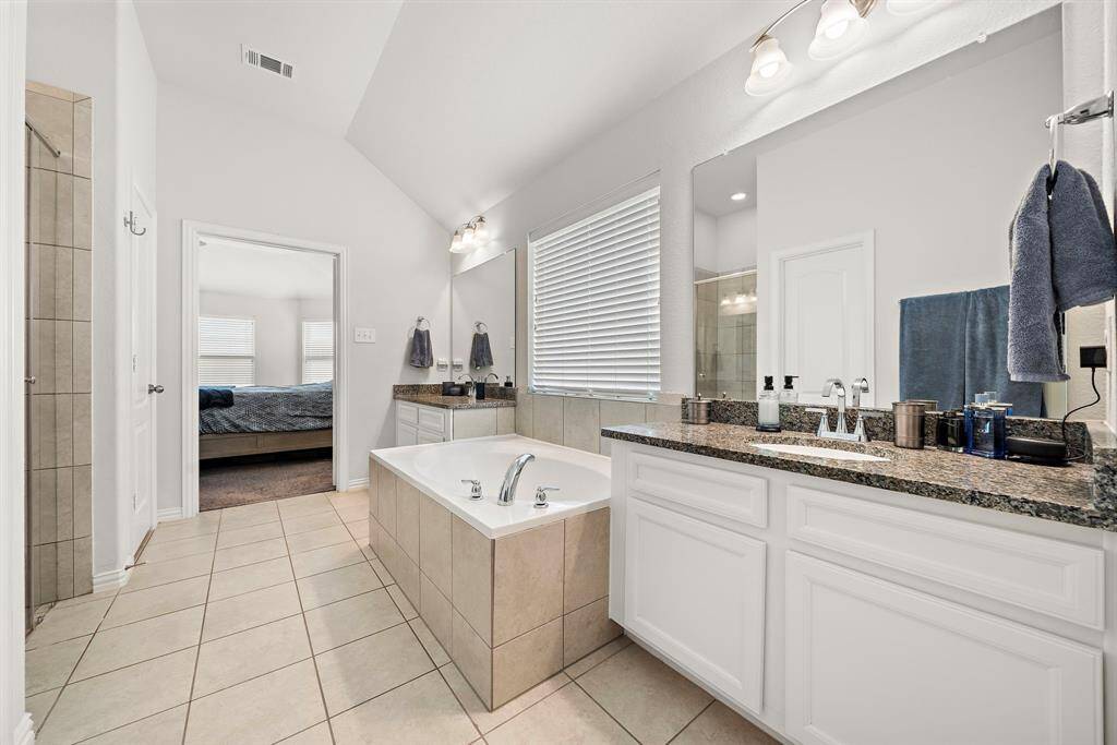 This spacious bathroom features a large soaking tub, dual sinks with granite countertops, and a separate shower. It connects to a bright bedroom, providing a convenient and luxurious setup.