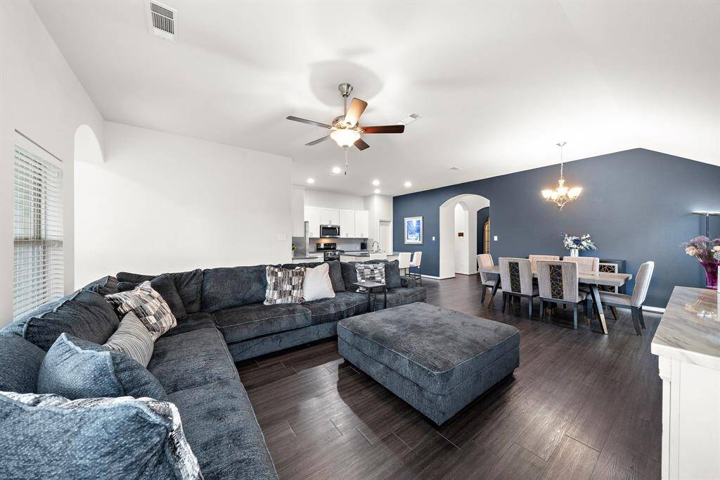 This spacious living and dining area features a modern open-concept design with dark hardwood floors. A ceiling fan and elegant lighting add comfort and style, while a deep blue accent wall enhances the dining space. The room is bright with natural light, offering a welcoming atmosphere.