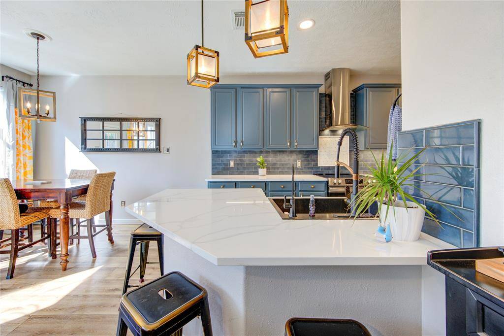 This modern kitchen features sleek blue cabinets, a stylish backsplash, and a spacious white countertop with a breakfast bar. Pendant lighting adds warmth, while natural light brightens the dining area with wicker chairs and a wooden table. Perfect for entertaining and everyday living.
