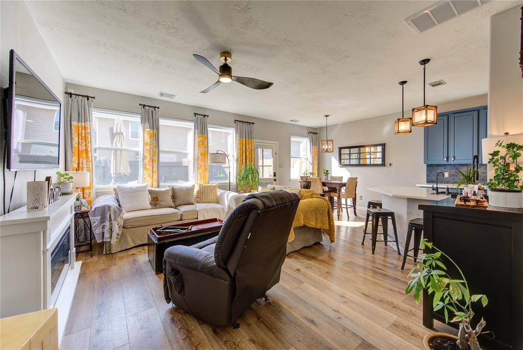 This inviting open-concept space features a well-lit living area with large windows, a cozy fireplace, and modern ceiling fan. The adjoining kitchen and dining area boasts a breakfast bar, stylish pendant lighting, and sleek cabinetry, all enhanced by warm wood flooring. Perfect for comfortable living and entertaining.