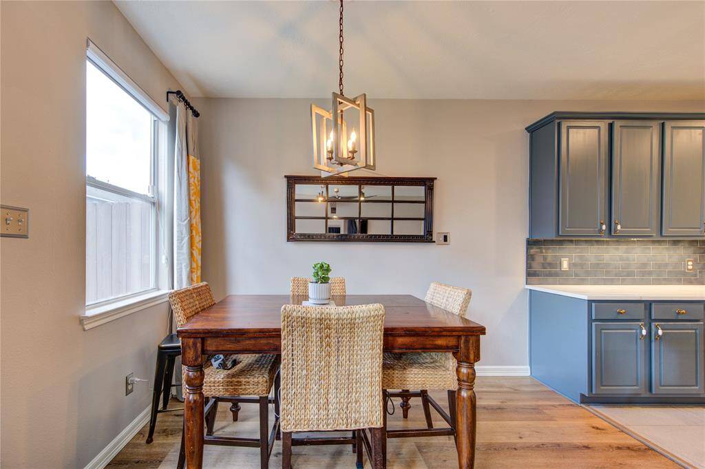 This photo showcases a bright dining area featuring a wooden table with wicker chairs, a stylish pendant light, and a large window. The adjacent kitchen has modern blue cabinetry and a sleek tile backsplash, creating a cozy and inviting space.