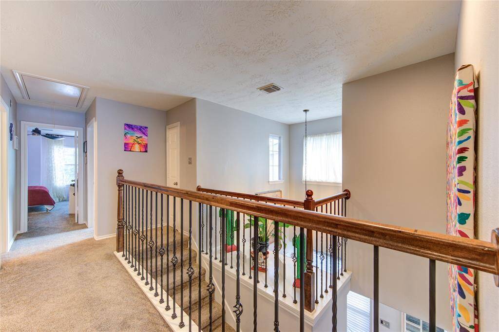 This photo features a bright and airy upstairs landing with carpeted floors and decorative iron railings. It leads to a bedroom and includes colorful wall art, adding a vibrant touch to the neutral walls. Natural light filters through a window, enhancing the open feel.