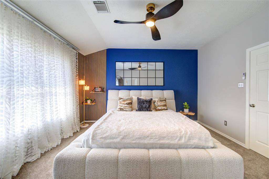 This photo features an upstairs hallway with carpeted flooring and decorative iron railings. It leads to a bright bedroom and a small window providing natural light. The walls are adorned with colorful artwork, adding a vibrant touch.