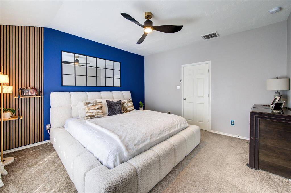 This bedroom features a cozy, modern design with a plush bed, a striking blue accent wall, and wooden slat detailing. It includes a ceiling fan, soft carpeting, and ample natural light, creating a relaxing and inviting atmosphere.