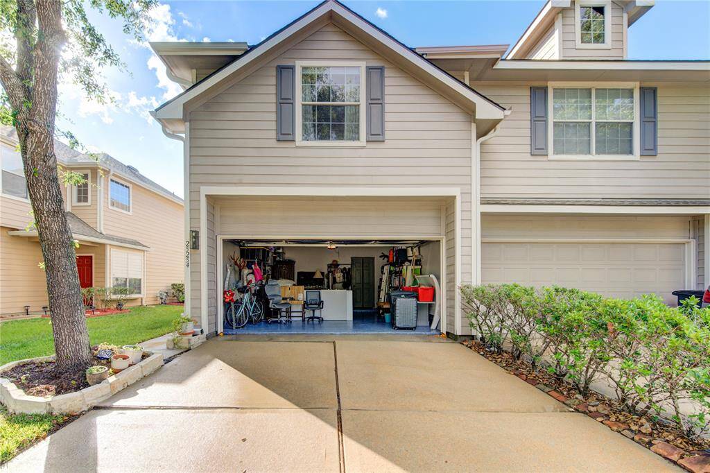 This photo shows a two-story house with a beige exterior and blue shutters. The open garage reveals ample storage and space, with a driveway leading up to it. The front yard is landscaped with a tree and shrubs, offering a welcoming curb appeal.