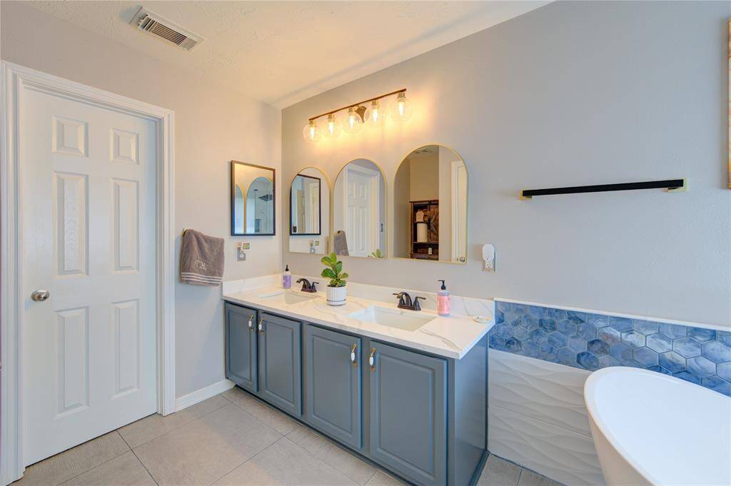This photo showcases a modern bathroom with dual sinks set in a sleek vanity, topped by stylish mirrors and contemporary lighting. The room features a soothing color palette, accentuated by a unique blue hexagonal tile backsplash and a freestanding tub, offering a luxurious and inviting atmosphere.
