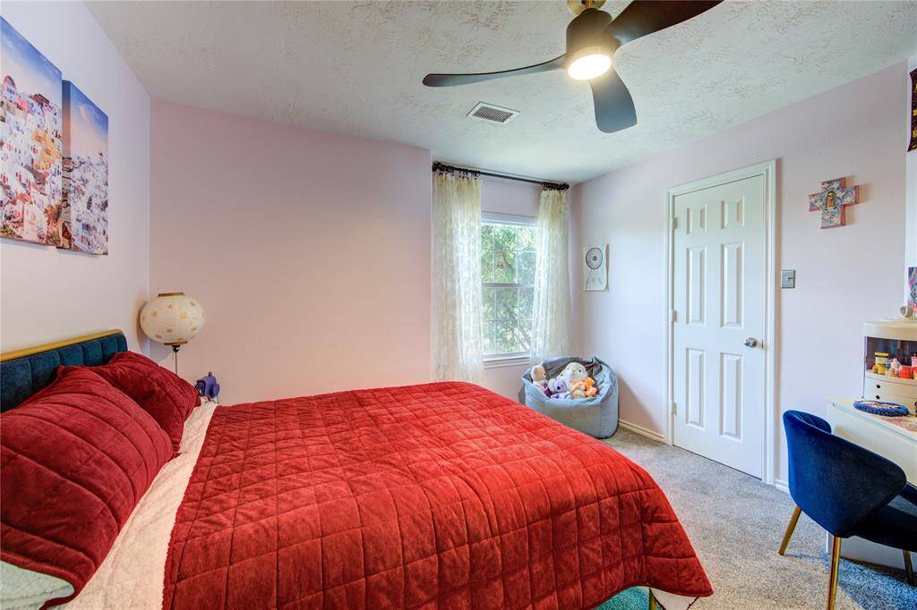 This cozy bedroom features a large window with natural light, a ceiling fan, and a plush red comforter on the bed. The room has soft carpeting, a desk with a chair, and a closet for storage, creating a comfortable and functional space.