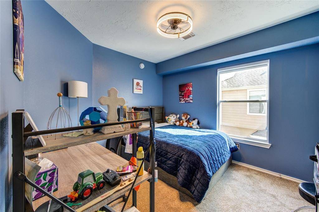Cozy bedroom with blue walls, carpeted floor, and a window providing natural light. Features a bed with plush toys, a modern ceiling light, and a small desk with shelves. Ideal for a child's room.