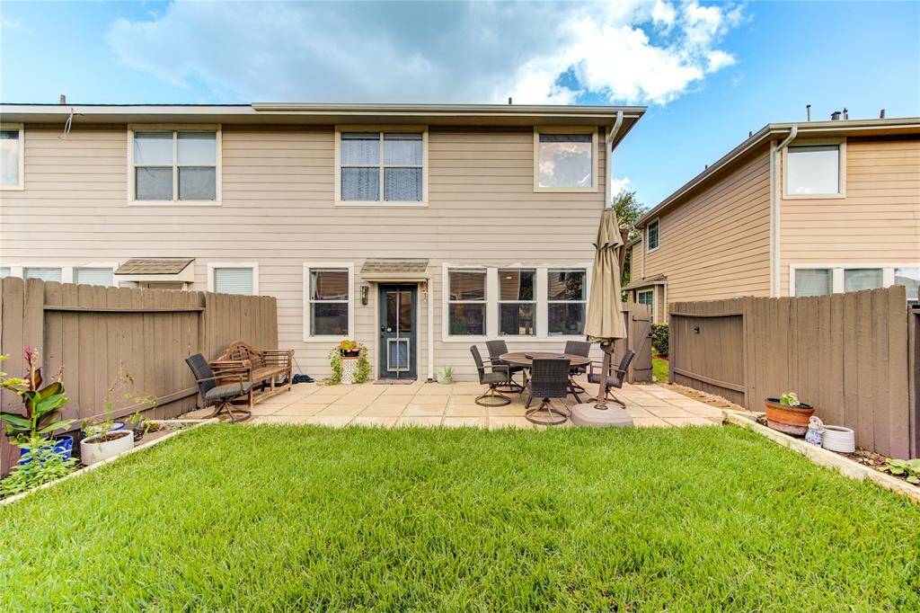 This photo shows a fenced backyard with a two-story house. The outdoor area features a patio with a dining set and umbrella, a small bench, and potted plants, offering a cozy space for relaxation and entertaining.