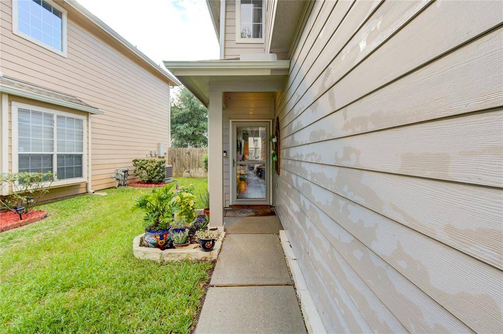 This photo shows a narrow entrance pathway leading to a home's front door, flanked by a well-maintained side yard with vibrant plants and clean siding. The space feels inviting and offers a glimpse of an adjacent house.