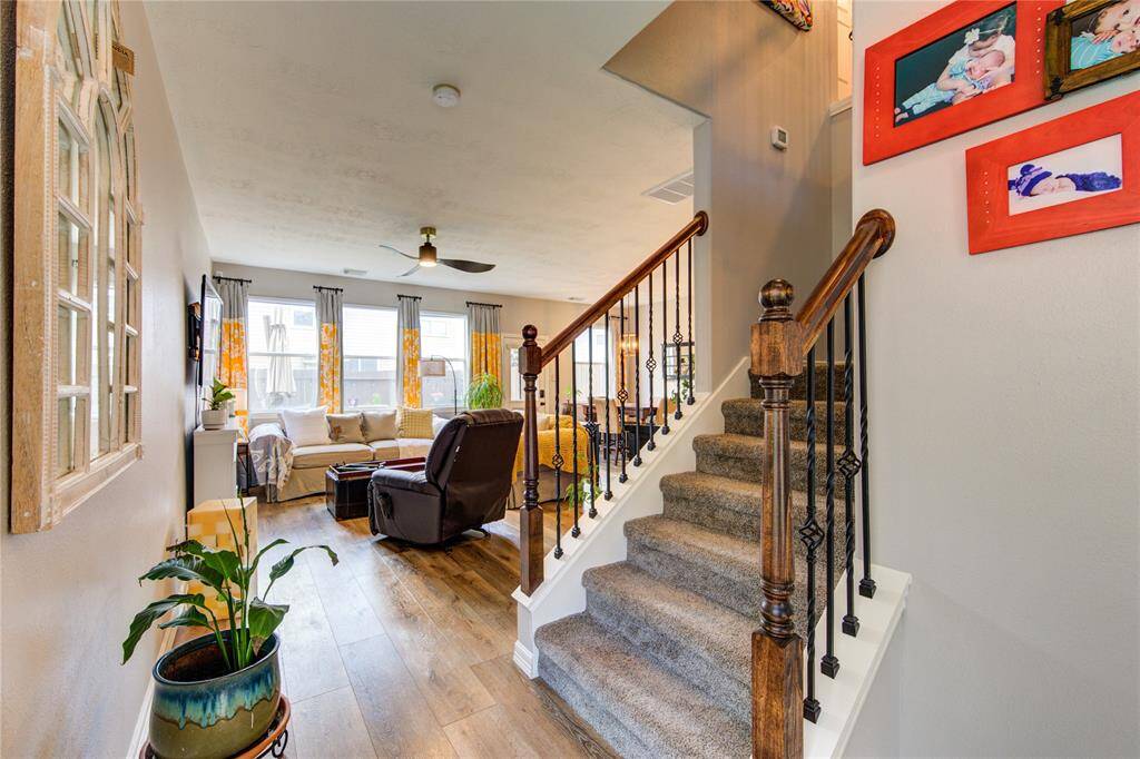 This inviting entryway features a welcoming front door with greenery, leading into a cozy living area. The space is bright with natural light, accentuated by wood flooring and a staircase with wrought iron railing. Decor adds a personal touch, creating a warm, homely atmosphere.