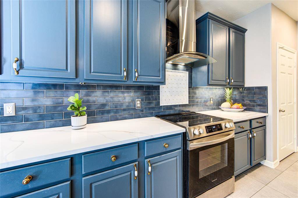 This kitchen features sleek blue cabinetry with brass hardware, a modern stainless steel oven, and a stylish blue tile backsplash. The white countertops provide a clean, bright look, complemented by a stainless steel range hood and tasteful decor.