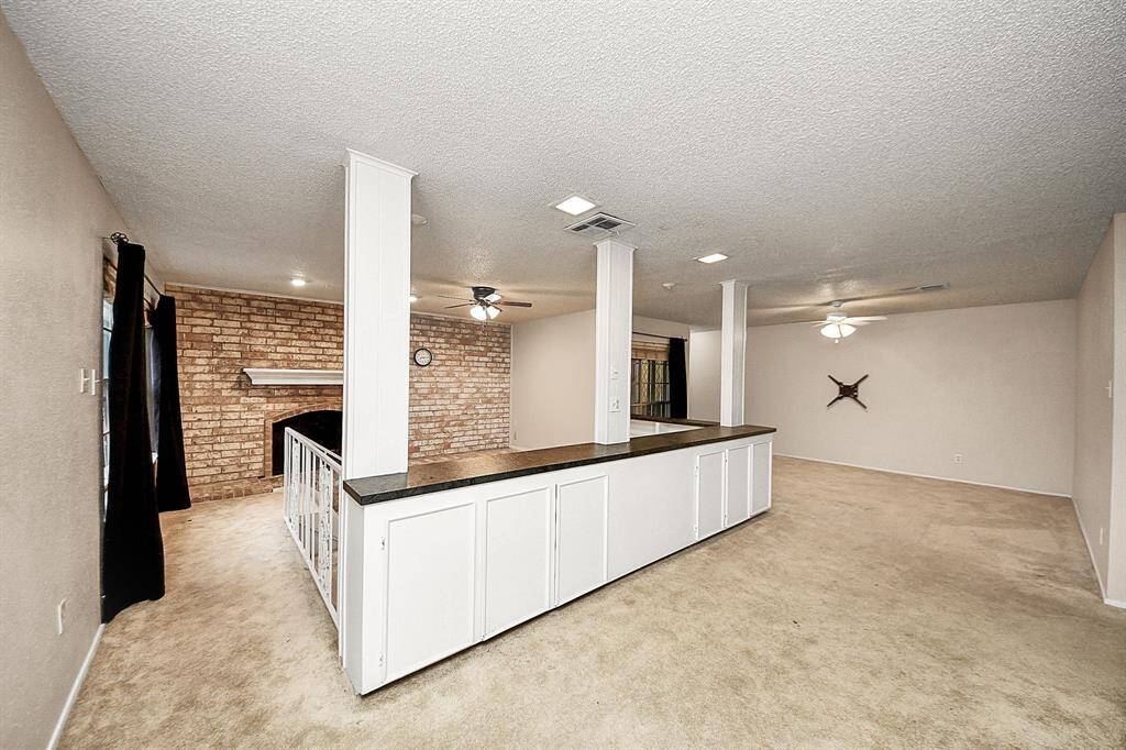 Large open-concept living space featuring neutral tones, built-in cabinetry, and multiple ceiling fans for comfort. The space flows seamlessly toward the brick fireplace and lower den area.