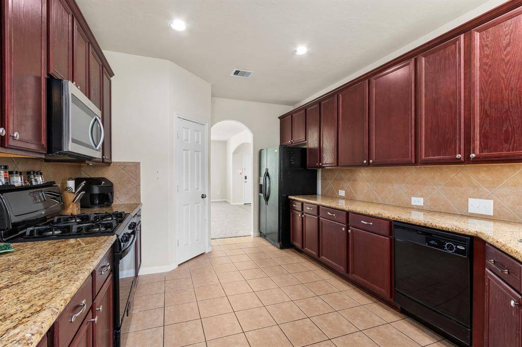 This kitchen features dark wood cabinetry, granite countertops, and stainless steel appliances. It has a spacious layout with tiled flooring and an arched entryway leading to another room.