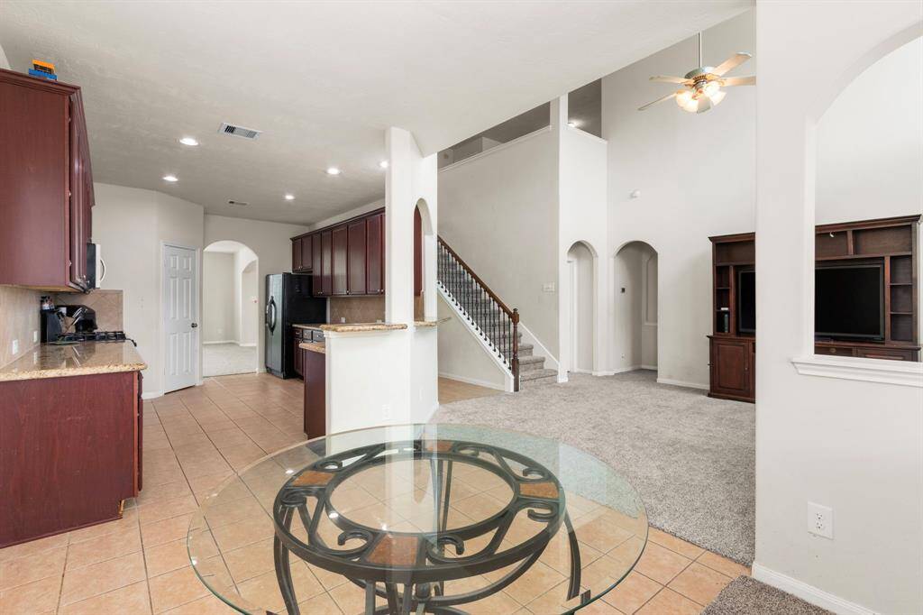 Spacious open-concept living area with high ceilings and a ceiling fan. The kitchen features dark wood cabinets, granite countertops, and tile flooring. A carpeted staircase leads to the upper level, and there is a built-in entertainment unit in the living room.