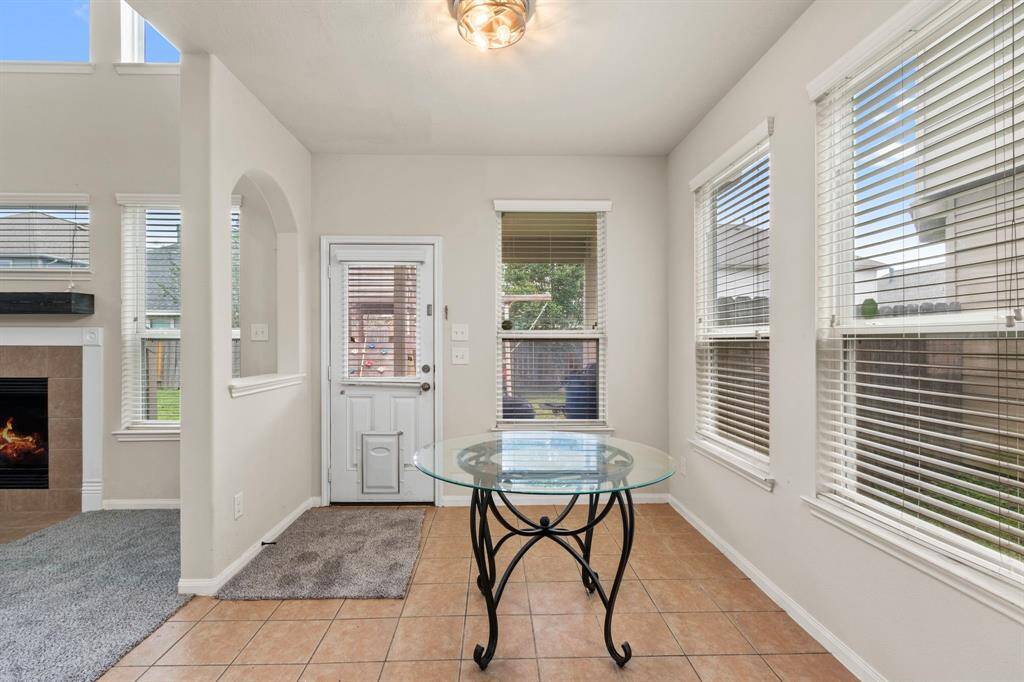 This photo showcases a bright, cozy space with tile flooring and ample natural light from multiple windows. It features a small glass table and a doorway leading to the outdoors, with a view of a fireplace in the adjacent carpeted room.