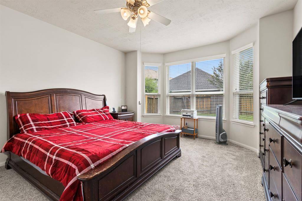 This bedroom features a cozy carpeted floor, a large window bay providing ample natural light, and a ceiling fan for comfort. It is furnished with a wooden bed frame and matching dresser, creating a warm and inviting atmosphere.