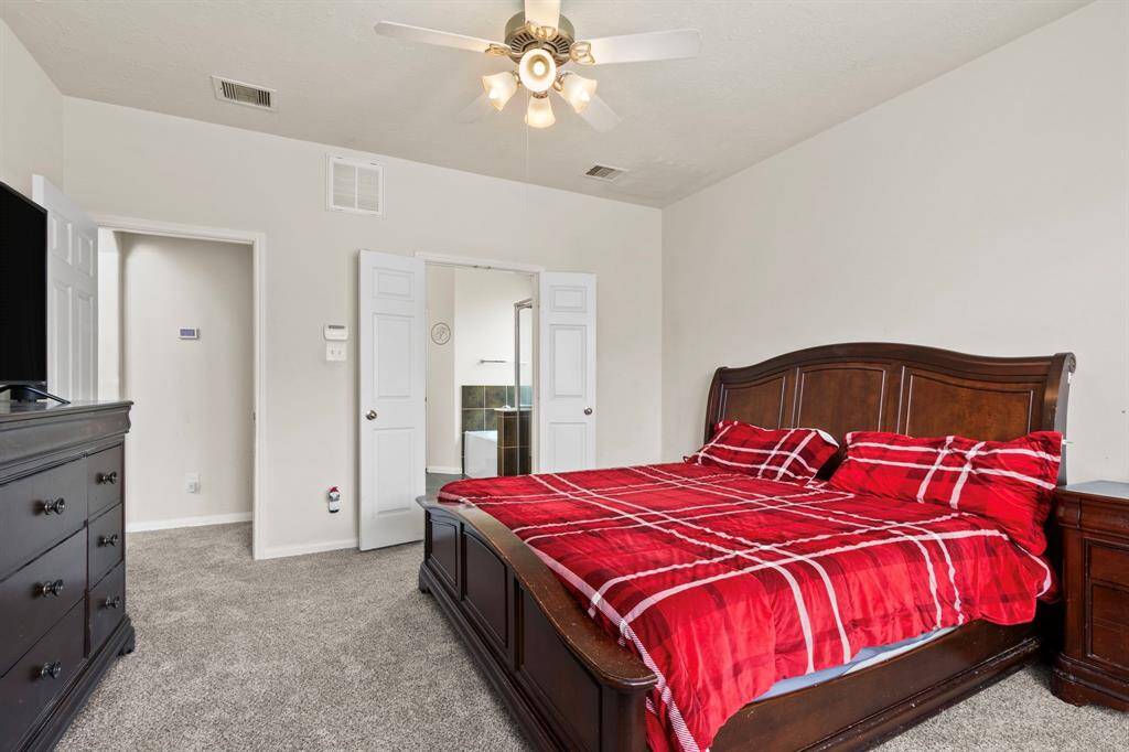 This spacious bedroom features a plush carpet, a ceiling fan, and elegant dark wood furniture. The room includes a large bed with vibrant red bedding and offers access to an en-suite bathroom through double doors.