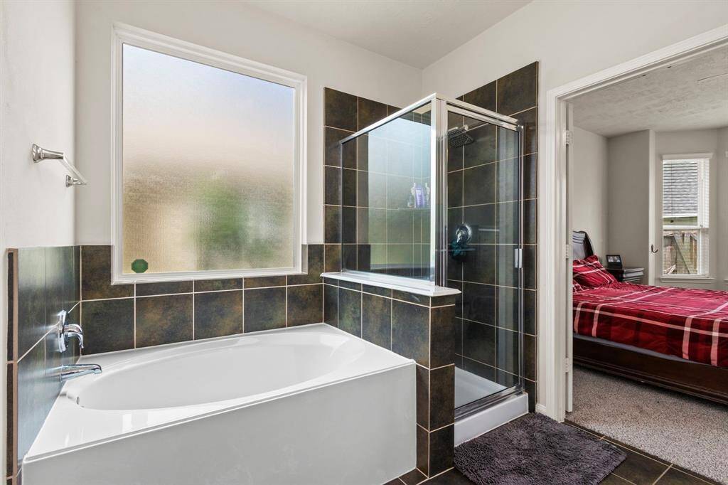 This bathroom features a large soaking tub and a separate glass-enclosed shower with dark tile accents. A frosted window provides natural light while maintaining privacy. The space connects to a bedroom with carpeted flooring.