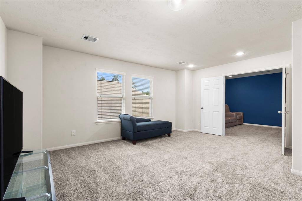 This photo showcases a spacious, carpeted living area with neutral walls, featuring a large window for natural light. It includes a cozy blue chaise and connects to an adjacent room through a doorway, offering a versatile layout.