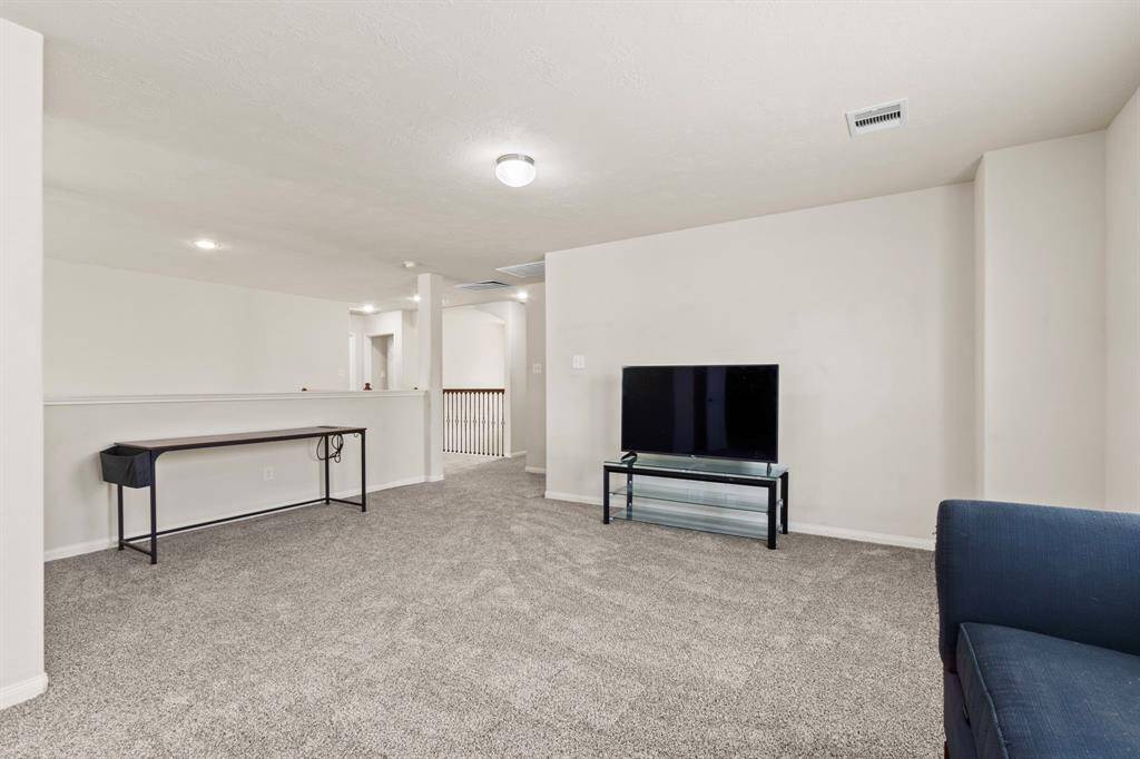 This room is a spacious, carpeted living area with neutral walls, featuring a TV setup and a small table. It has ample lighting and an open layout leading to a hallway, ideal for relaxation and entertainment.