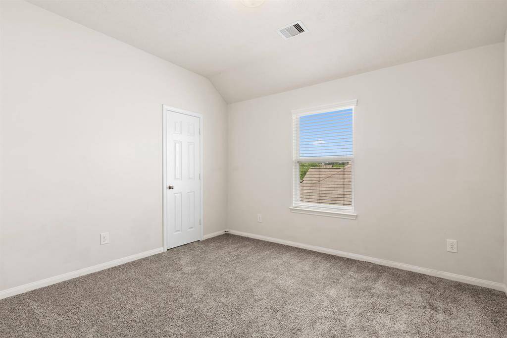 This is a small, bright bedroom with neutral walls and carpet. It features a single window with blinds, offering natural light, and a standard white door.