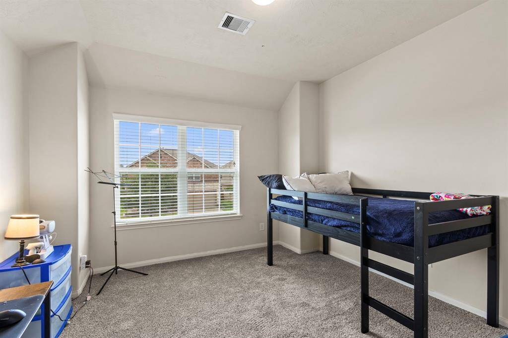 This is a cozy, carpeted bedroom with neutral walls, featuring a window that allows ample natural light. It includes a loft bed and a small desk area, perfect for a child's room or a compact office space.