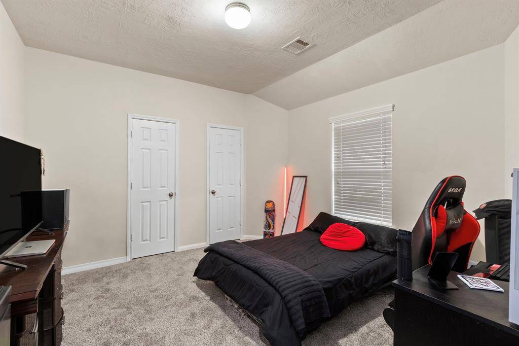 This room features a cozy space with neutral walls and carpeted flooring. It includes a bed, a gaming chair, and a desk setup, making it versatile for both relaxation and work. Two closets offer ample storage, and a window provides natural light.